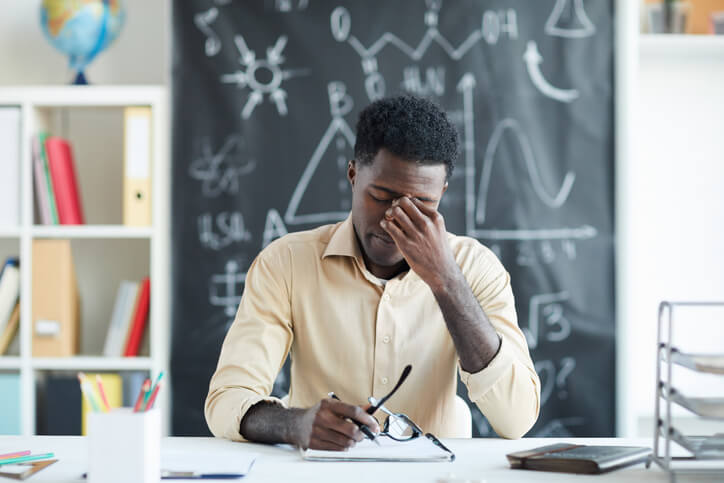 Tired young teacher sitting by desk in classroom and touching nose bridge in front of chalkboard