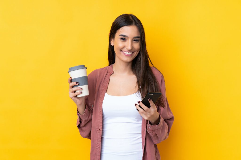 brunette girl holding coffee and using cell phone