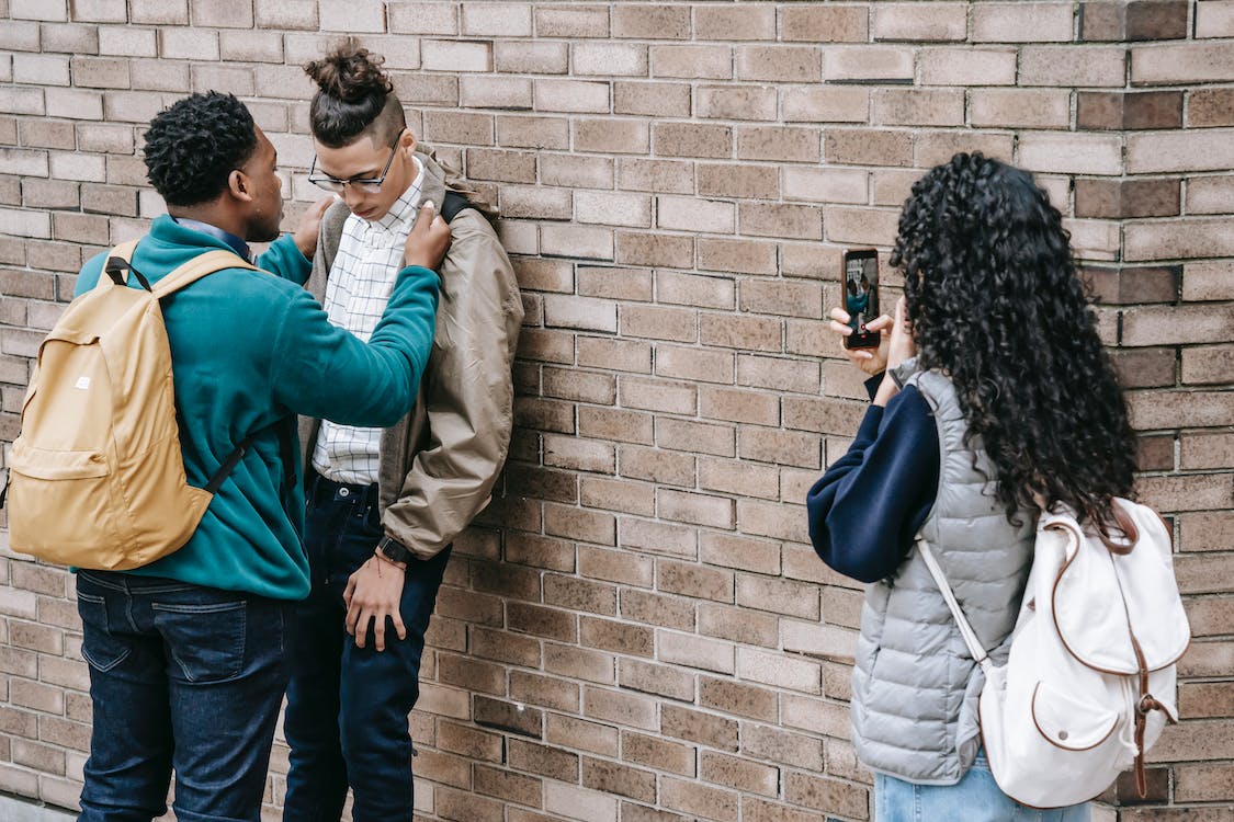 Student bullies another pushing against brick wall while other takes pics with phone