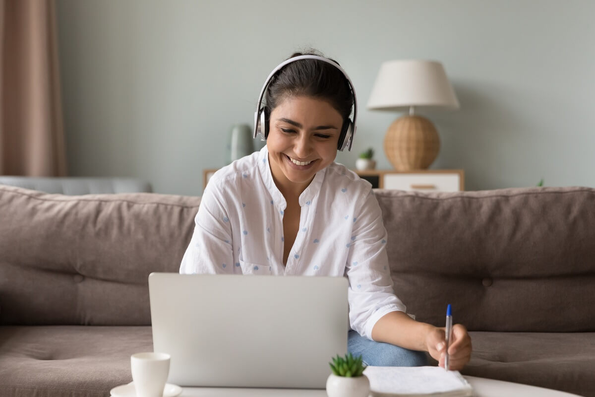 Cheerful Indian woman educator in headphones studying at home seated on sofa at table with laptop