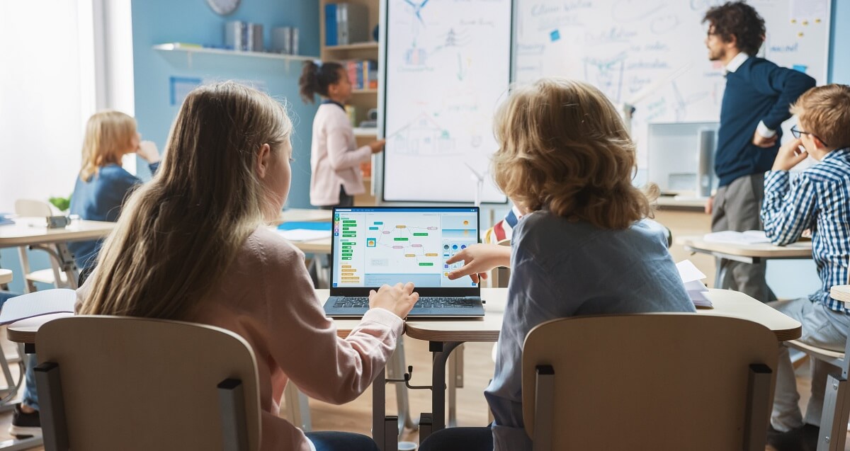 Classroom over shoulder of little boy and girl using programming software on laptop while teacher explains lesson