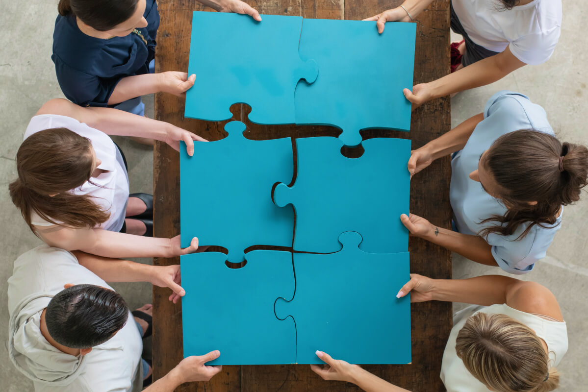 overhead view of six coworkers holding together completed blue 6 piece hexagon puzzle over table