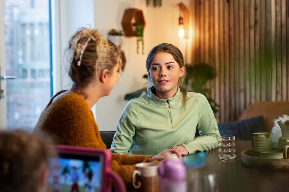 Mother talking to female tween at kitchen table and sibling on tablet
