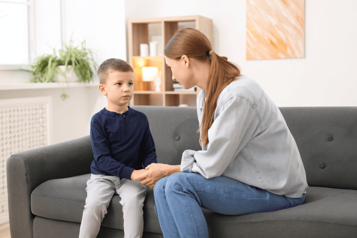 Mother sitting on a sofa holding a young boy’s hands while talking to him calmly at home