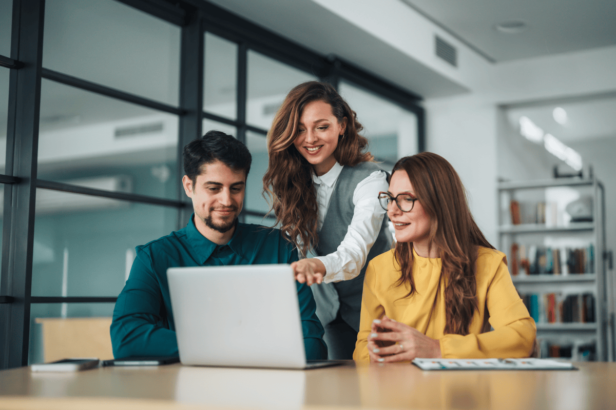 At a table, a BCBA stands between two seated colleagues at their ABA clinic and points to a laptop running an ABA practice management platform